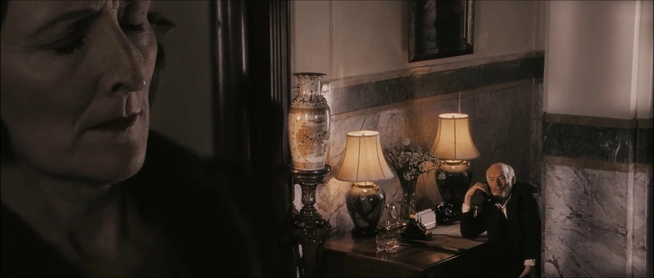 The Black Dahlia (2006) movie still: closeup — A dark hallway with marble walls contains a wooden table with lamps and a vase. An elderly man sits…