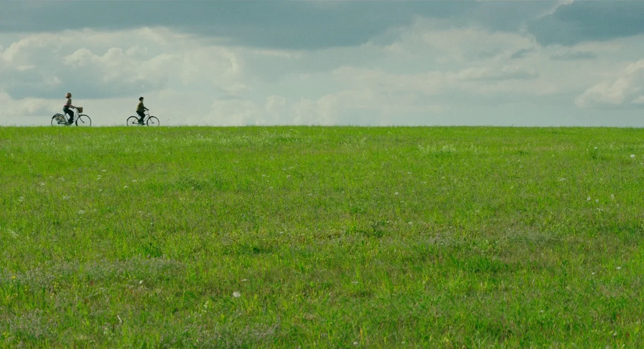 Jojo Rabbit (2019) movie still: wide — A vast, open green meadow stretches toward the horizon under a heavily clouded sky. Two individuals on…