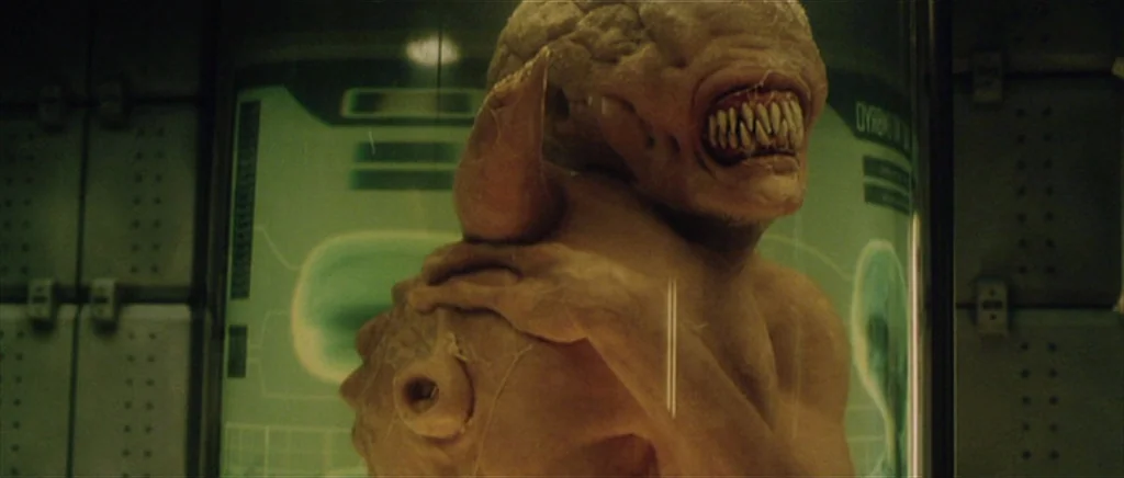 Alien: Resurrection (1997) movie still: closeup — A biological specimen is encased within a glowing glass cylinder in a dark laboratory. The…