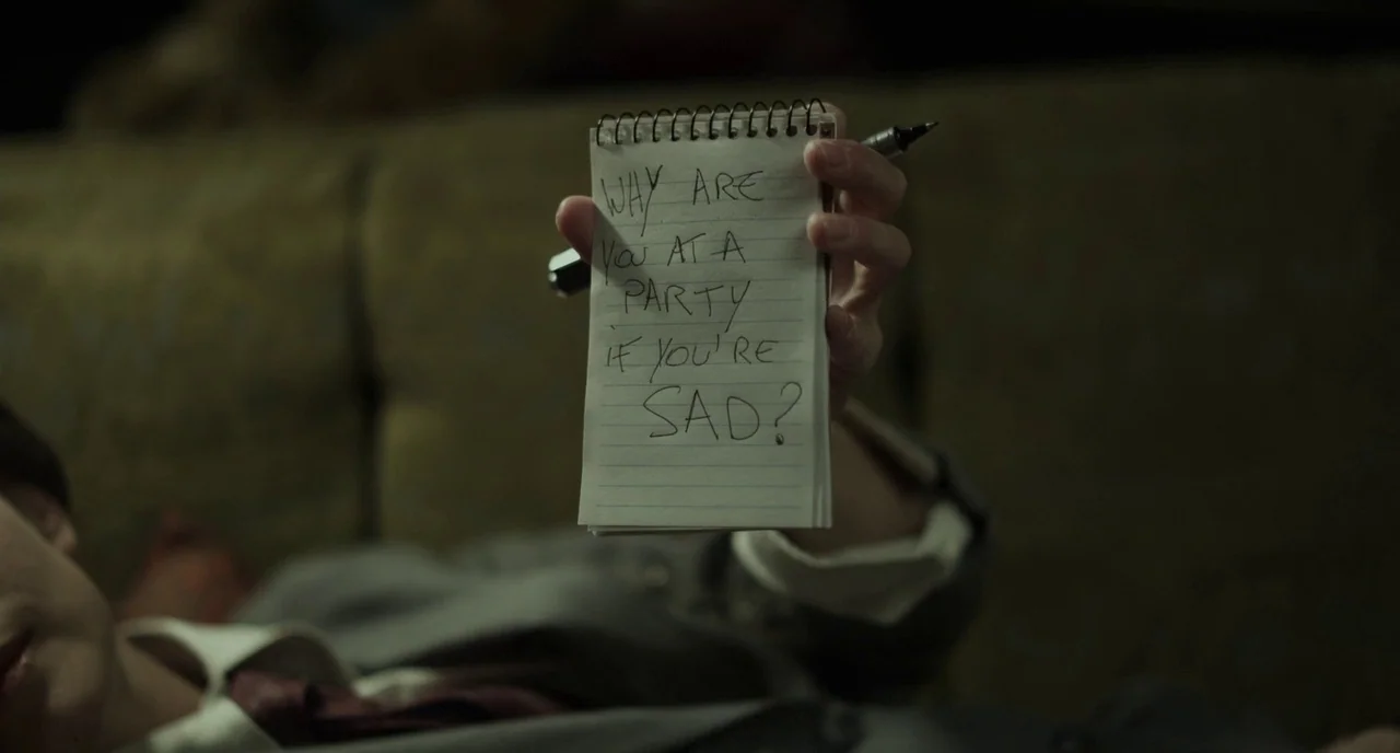 Beginners (2010) movie still: closeup — A small spiral notepad is held up against a blurry, dark green sofa background. The paper contains the…
