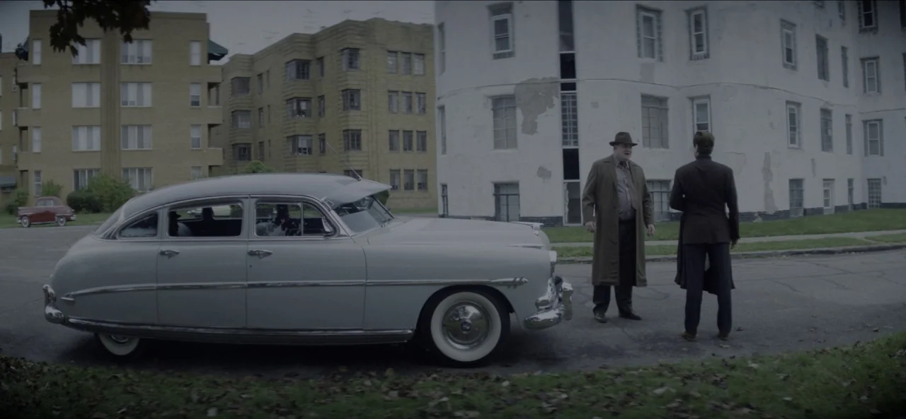 No Sudden Move (2021) movie still: wide — A residential street lined with multi-story apartment buildings features a parked 1950s automobile. Two…