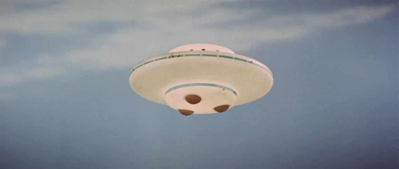Invasion of Astro Monster (1965) movie still: wide — A pale circular disc-shaped aircraft hovers against a plain light blue overcast sky. The object…