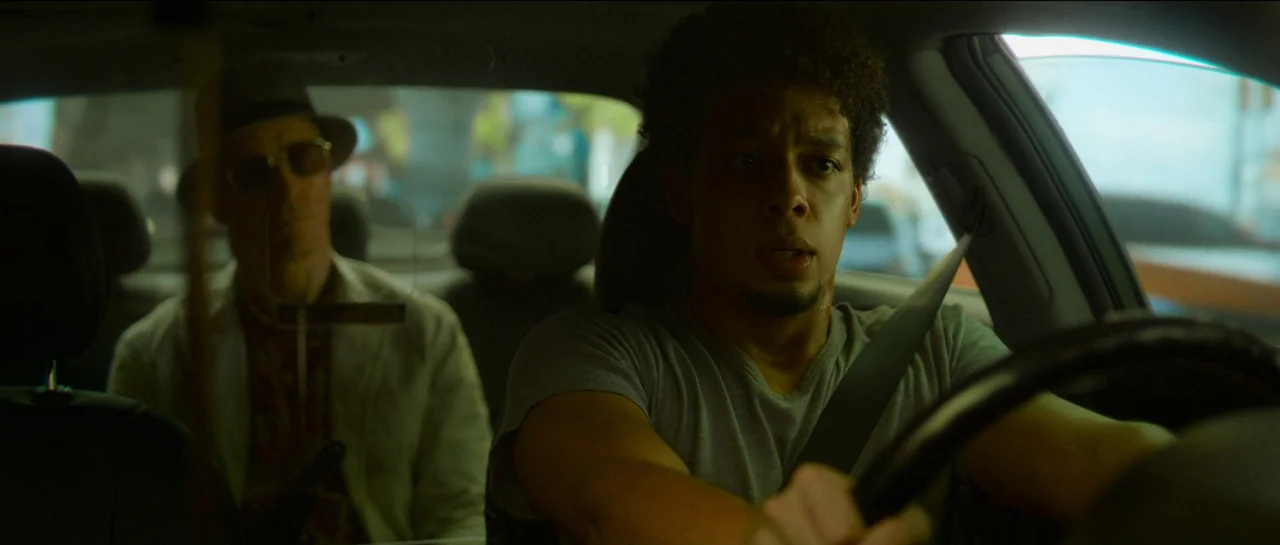 The Killer (2023) movie still: medium — A man drives a car during the day with a passenger in the back seat. Outside the windows, street life is…