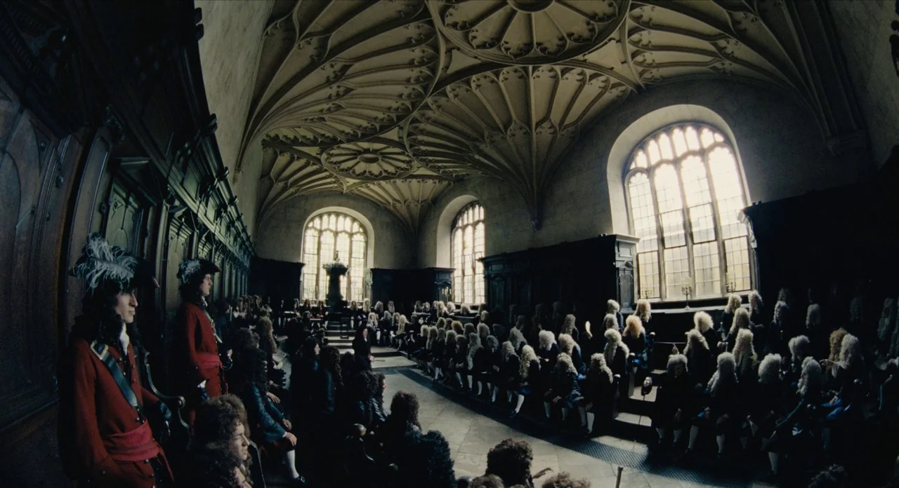 The Favourite (2018) movie still: wide — A historic chamber with vaulted ceiling features rows of seating in tiered wooden benches. A large group of…