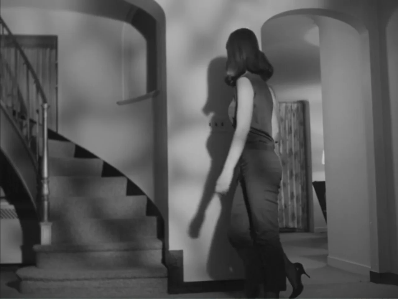 Chained Girls (1965) movie still: fullbody — A residential foyer contains a curved staircase with a dark wooden banister. A woman walks toward the…