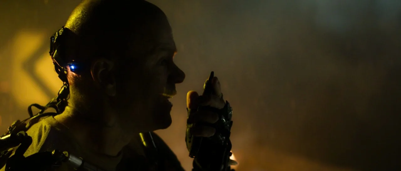 Elysium (2013) movie still: closeup — A dark industrial space filled with dim, yellow-hued atmosphere and mechanical clutter. A bald man wearing a…