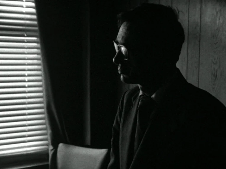 Death and Transfiguration (1983) movie still: medium — A dimly lit interior space with wood-paneled walls and a window with closed blinds. A man in…