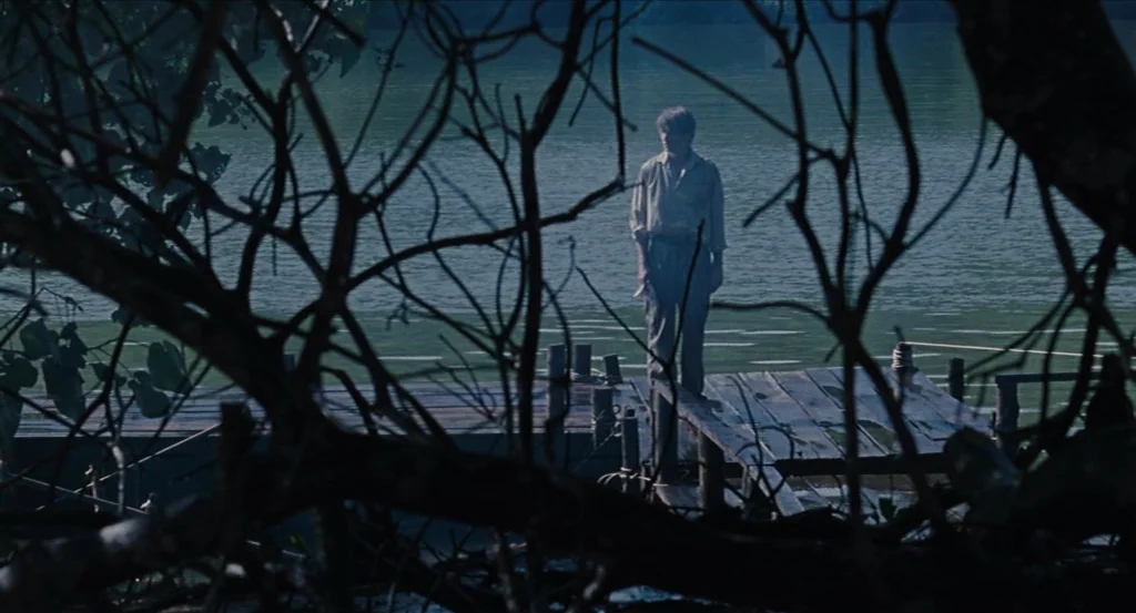 Almayer’s Folly (2011) movie still: wide — A wooden pier extends into a wide, calm lake surrounded by dense, dark vegetation. A solitary man stands…
