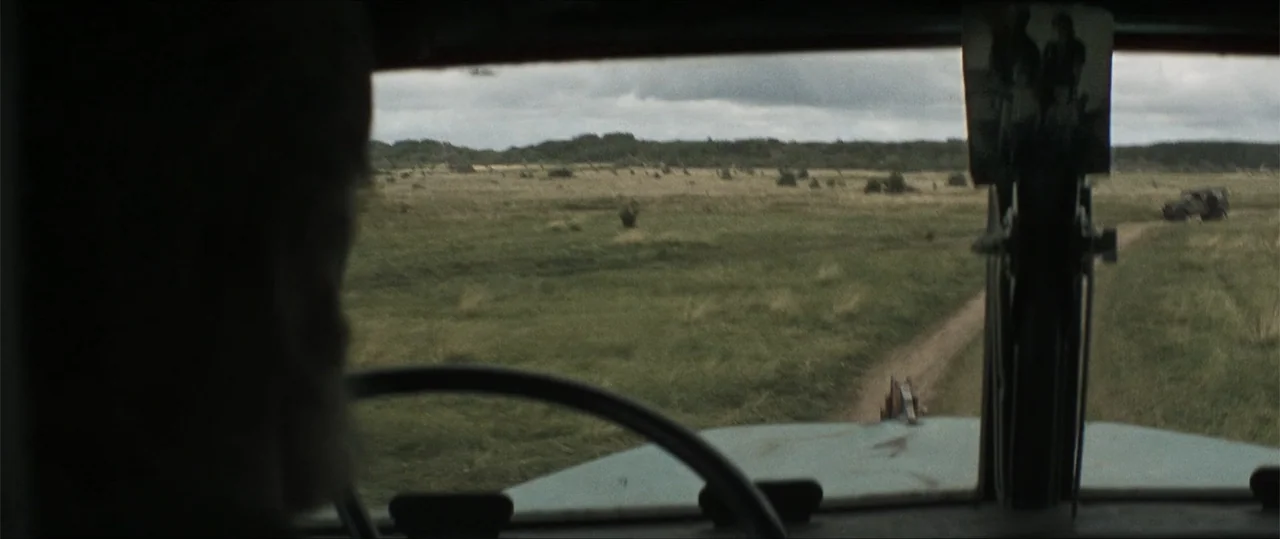Sisu: Road to Revenge (2025) movie still: medium — The view from inside a vehicle looking out across an expansive grassy rural field. A narrow dirt…