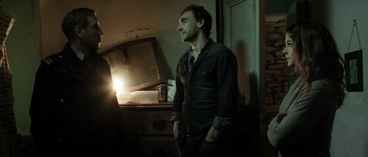 The Hallow (2015) movie still: medium — A dimly lit room with peeling walls contains three people standing in a group. They face one another near a…