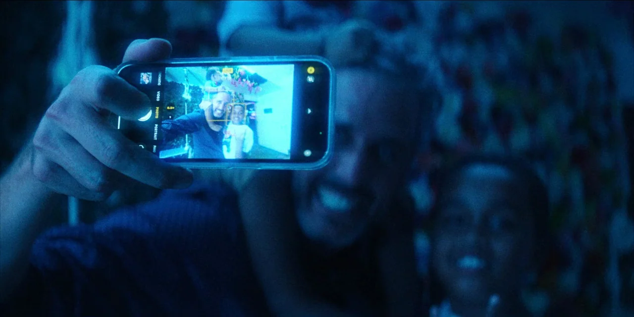Spermworld (2024) movie still: closeup — A man holds a smartphone in a dimly lit, blue-tinted environment. The screen illuminates his face and the…