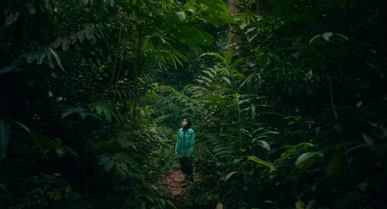 Tiger Stripes (2023) movie still: wide — A dense, overgrown tropical forest with tall trees and thick green ferns on all sides. A young person…