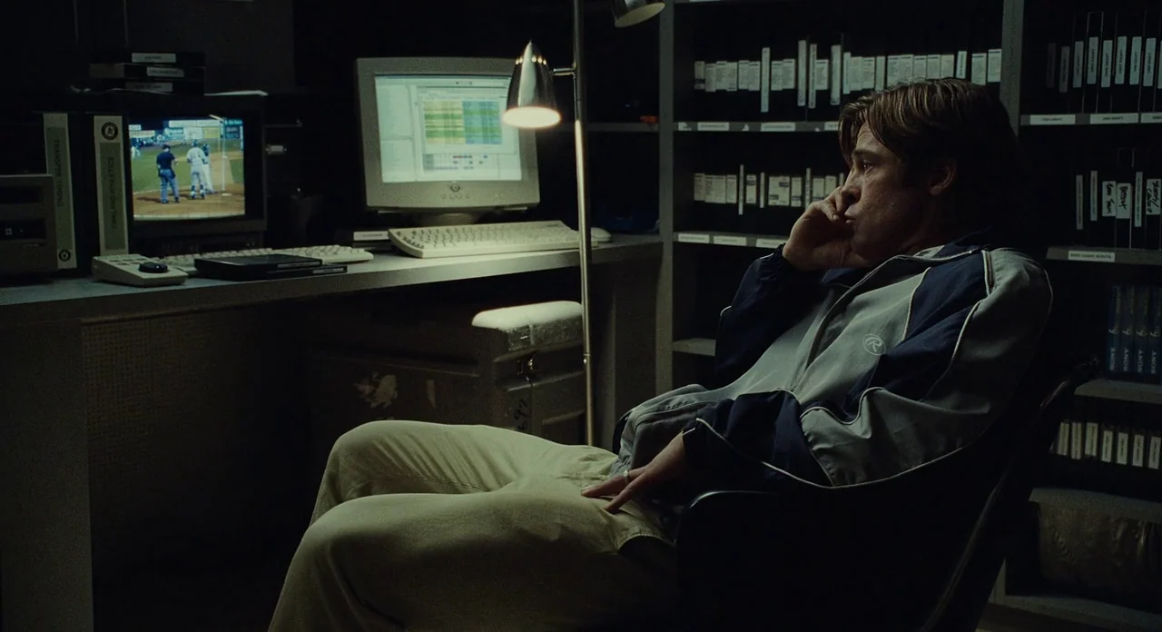 Moneyball (2011) movie still: medium — A cramped, dark workspace filled with shelves of video tapes and computer hardware. A man sits at a desk…