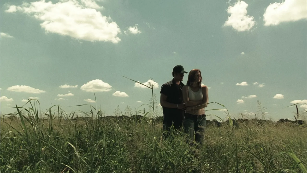 The Texas Chainsaw Massacre (2003) movie still: fullbody — An open grassy field with scattered clouds in a pale blue sky. A man and a woman stand…