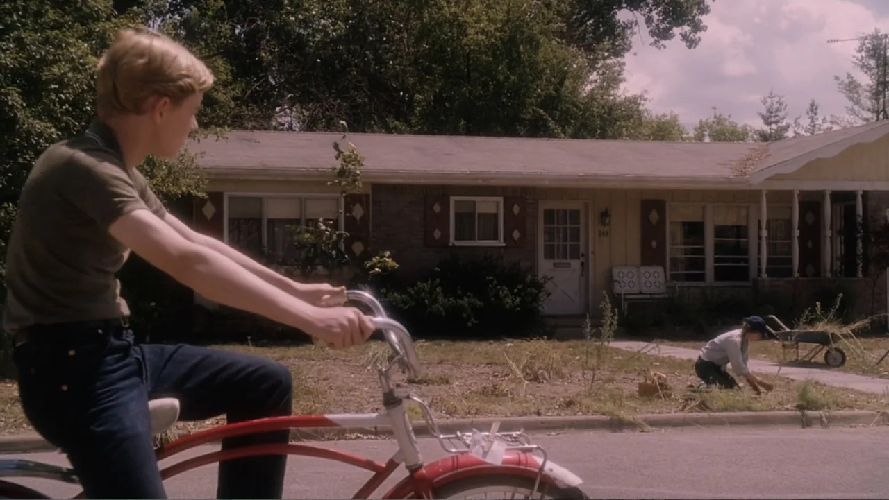 Flipped (2010) movie still: medium — A residential street in front of a single-story house with a small lawn. A boy on a bicycle pauses in the…