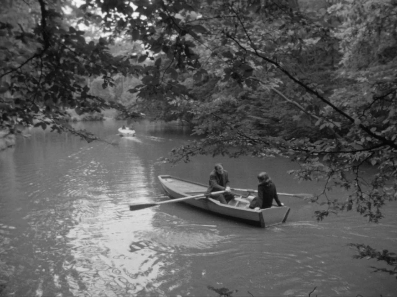 A Woman Kills (1968) movie still: wide — A calm lake surrounded by dense forest trees overhangs the water surface. A man and woman sit in a small…