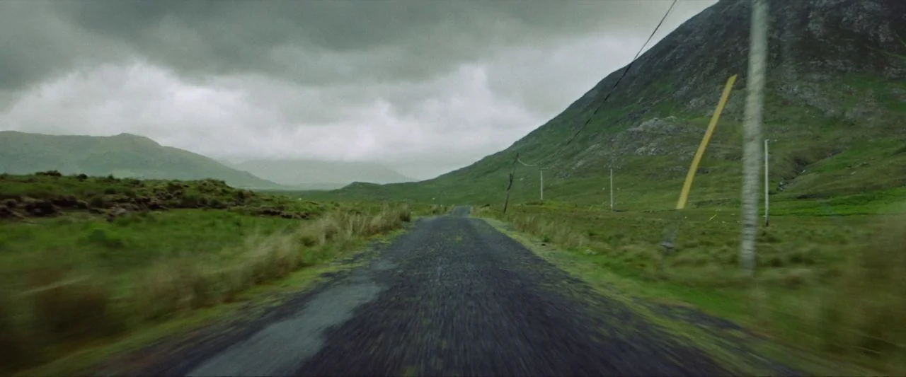 Calm With Horses (2019) movie still: wide — A wet asphalt road winds through a vast, green, mountainous valley under heavy clouds. Telephone poles…