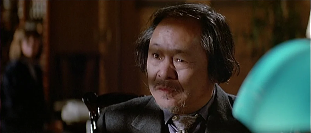 Big Trouble in Little China (1986) movie still: medium — A dimly lit room contains wooden furniture and a visible green desk lamp. An elderly Asian…