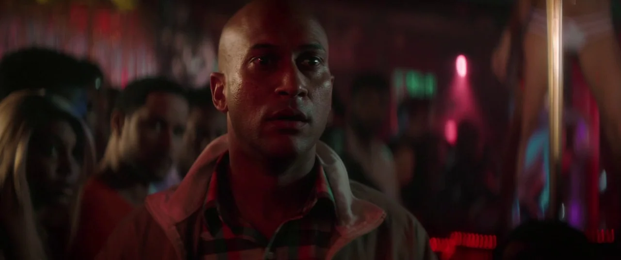 Keanu (2016) movie still: medium — A dark, crowded room features neon lighting cast across the faces of patrons. A bald man stands in the foreground…