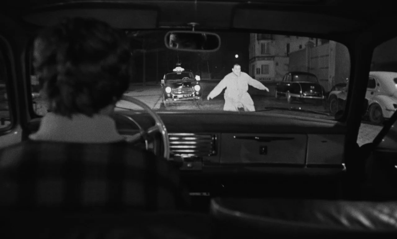 Witness In The City (1959) movie still: medium — A dark night street is illuminated by car headlights in a city environment. A man in light clothing…