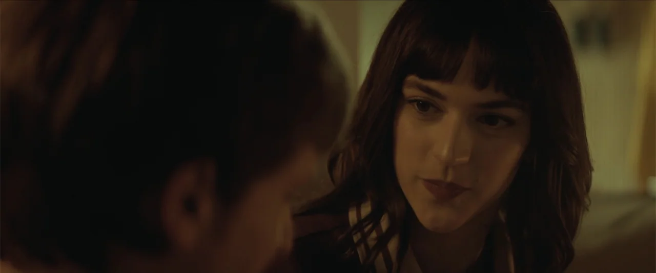 Tell Me Softly (2025) movie still: closeup — A dimly lit room provides a background for two people in conversation. A woman stares at a man seen…