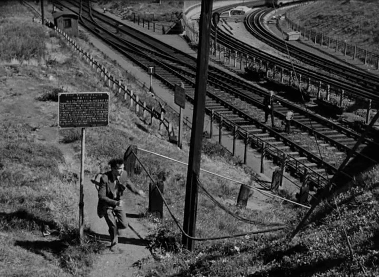 The Blue Lamp (1950) movie still: wide — A steep, grassy railway embankment features a sign and complex overhead utility wires. A man in a dark suit…
