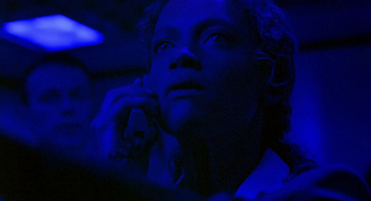 Resident Evil (2002) movie still: closeup — An interior space lit by a bright blue overhead light fixture at night. A woman in the foreground holds…