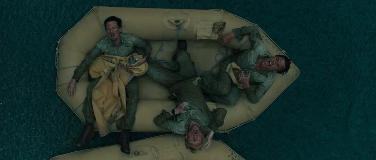 Unbroken (2014) movie still: wide — Three soldiers sit in a tan inflatable life raft floating on dark ocean water. The men are exhausted and appear…