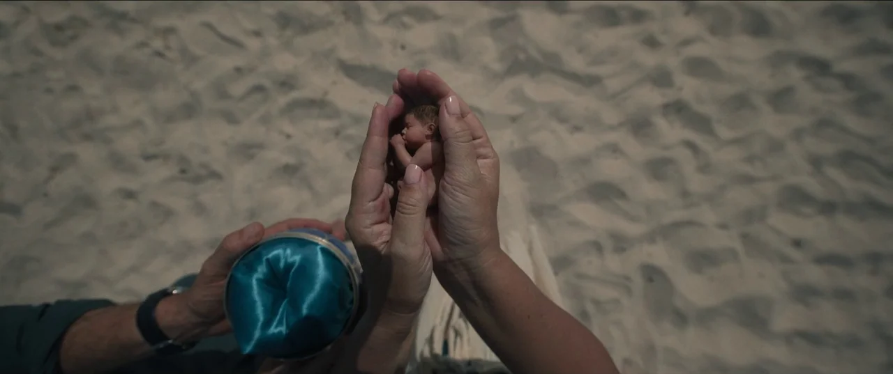 Bardo: False Chronicle of a Handful of Truths (2022) movie still: closeup — A person's hands cup a tiny, realistic infant figure over a textured…