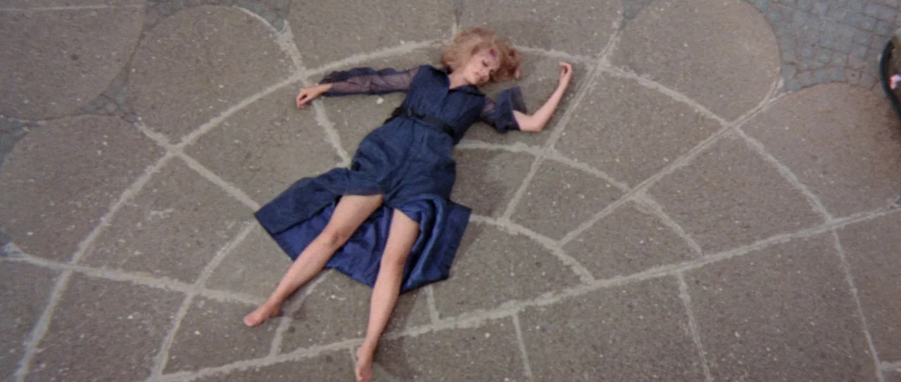 Orgasmo (1969) movie still: fullbody — A stone-paved plaza features a fan-shaped pattern laid out in grey paving stones. A young woman in a navy…