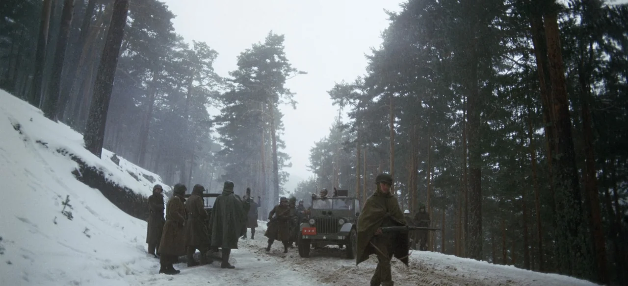 Patton (1970) movie still: wide — A group of soldiers stands on a snowy road flanked by dense pine trees. A military jeep is parked in the middle of…