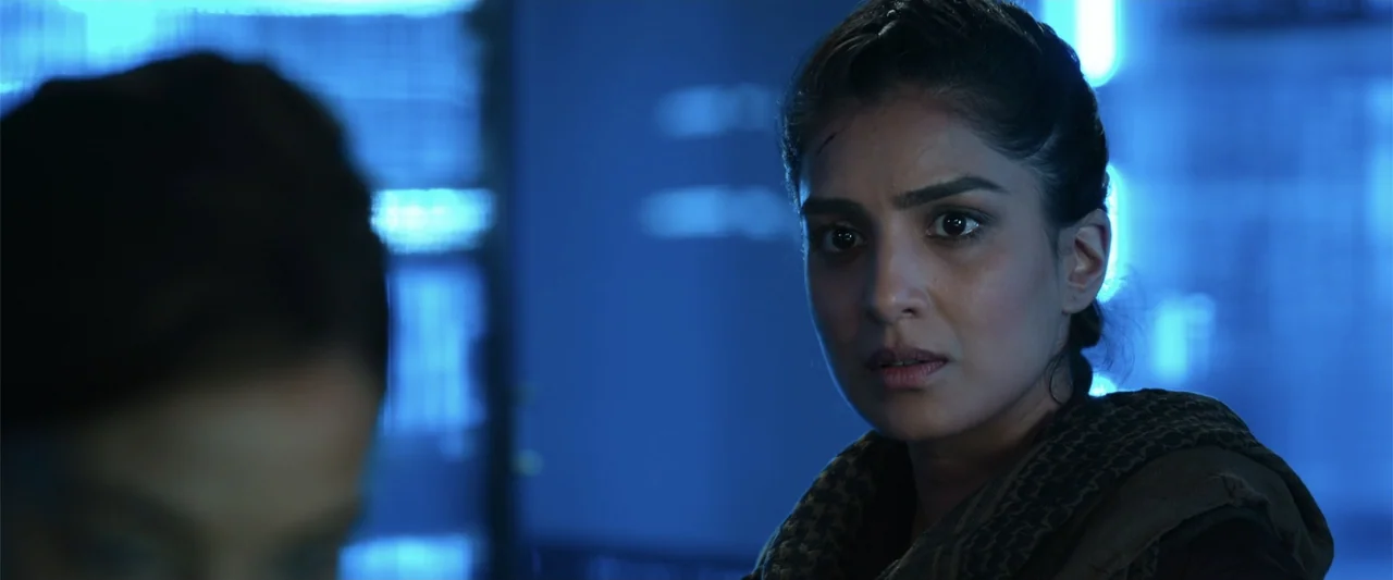 Black Site (2022) movie still: closeup — A dimly lit room filled with blue computer hardware illumination. A woman with a forehead cut looks…