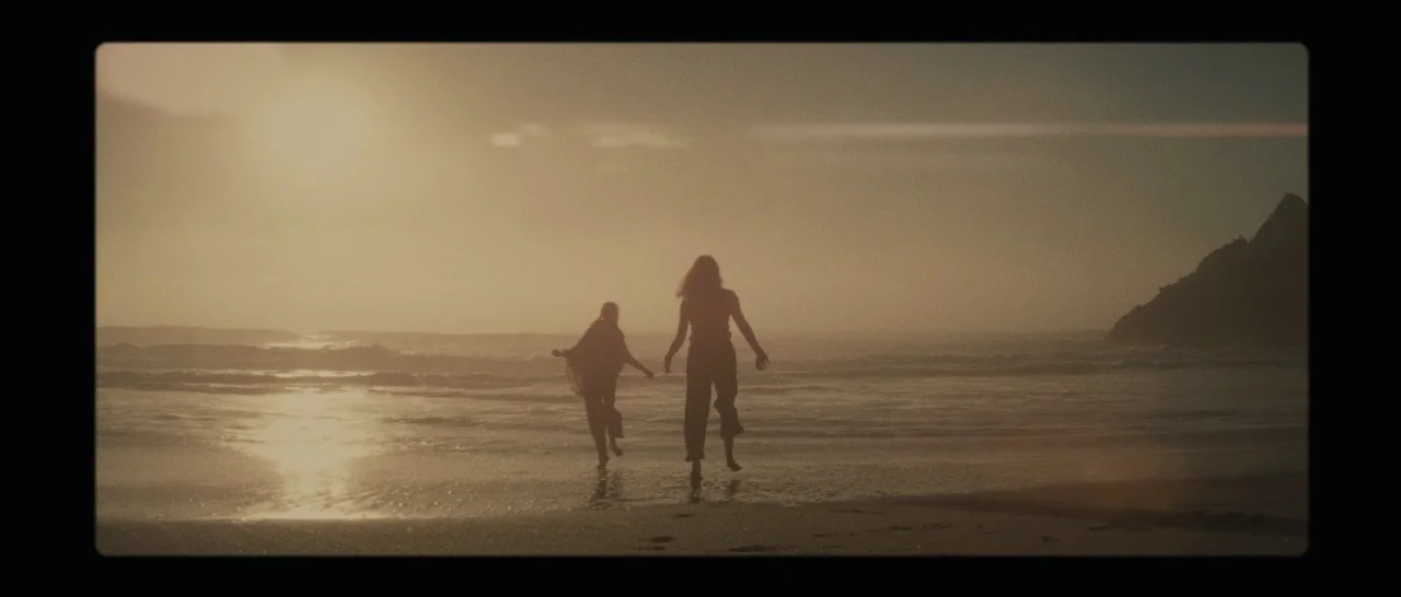 The Diary of a Teenage Girl (2015) movie still: wide — A wet sand beach stretches toward the ocean at sunset with a rocky cliff. Two people run into…