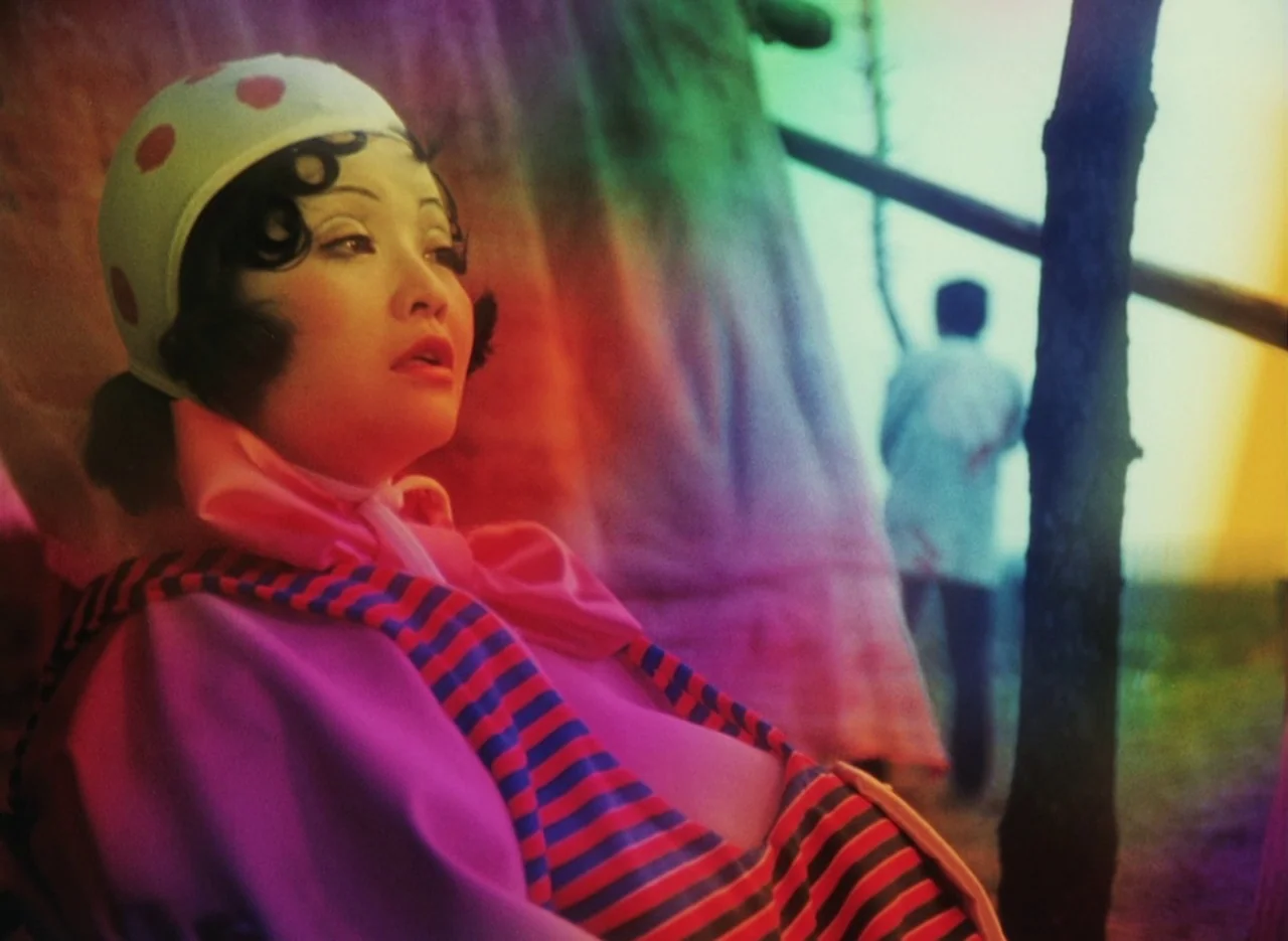 Pastoral To Die In The Country (1974) movie still: closeup — A woman wearing a spotted cap sits outdoors with a hazy landscape behind her. A blurred…