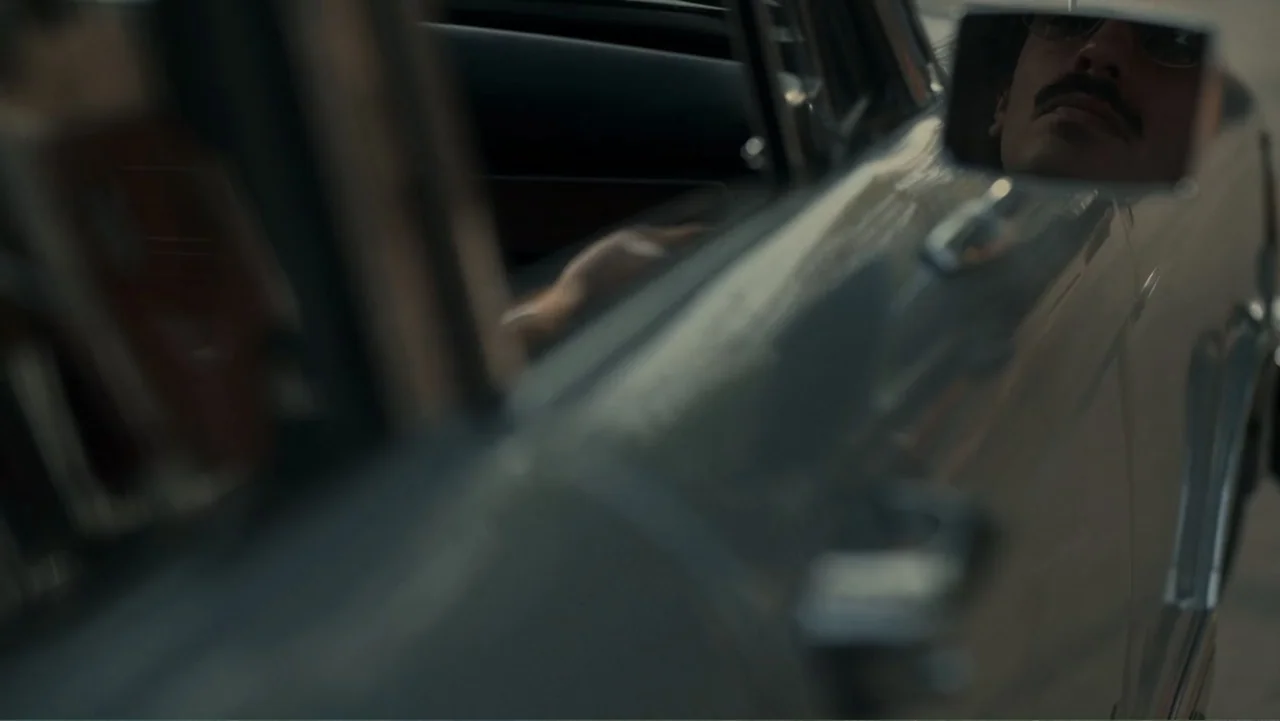 Transfers (2024) movie still: closeup — A close-up view shows a car side-view mirror reflecting a man's face. The car's metal frame is in focus…
