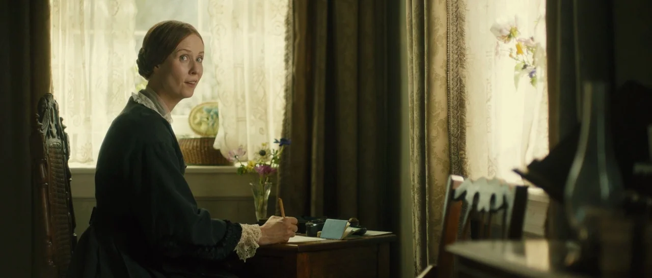 A Quiet Passion (2016) movie still: medium — A 19th century room with patterned curtains and a wooden desk near a window. A woman in a dark dress…