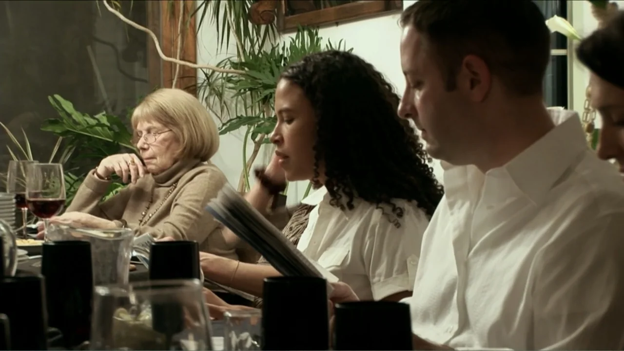 Little White Lie (2008) movie still: medium — A group of people sits at a dining table during an evening event indoors. Three individuals are…