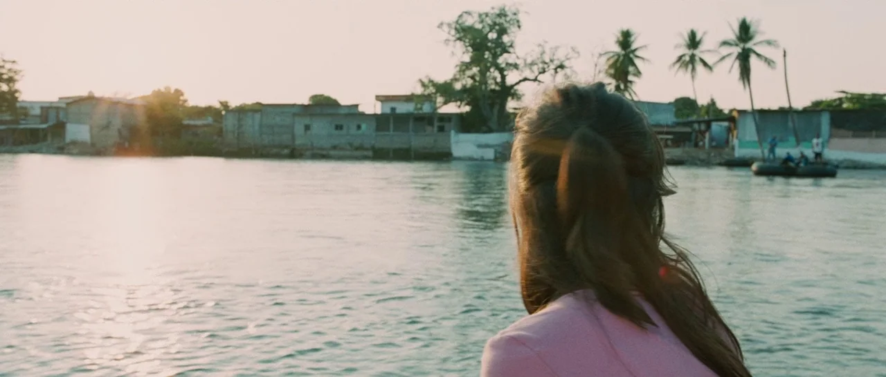 Sin Nombre (2009) movie still: medium — A person looks toward a shoreline with small buildings and palm trees across water. The sun sets low on the…