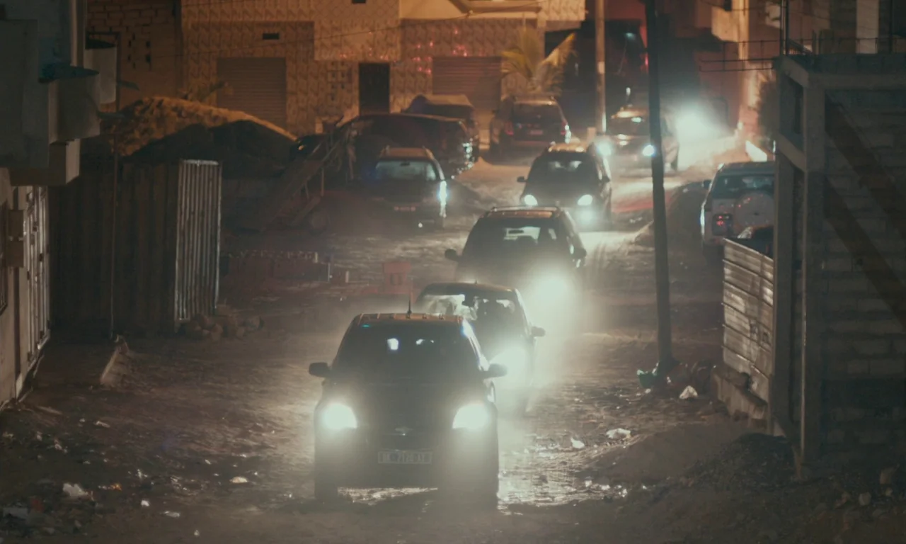 Atlantics (2019) movie still: wide — A narrow dirt road at night is crowded with several cars driving forward. Dust particles are suspended in the…