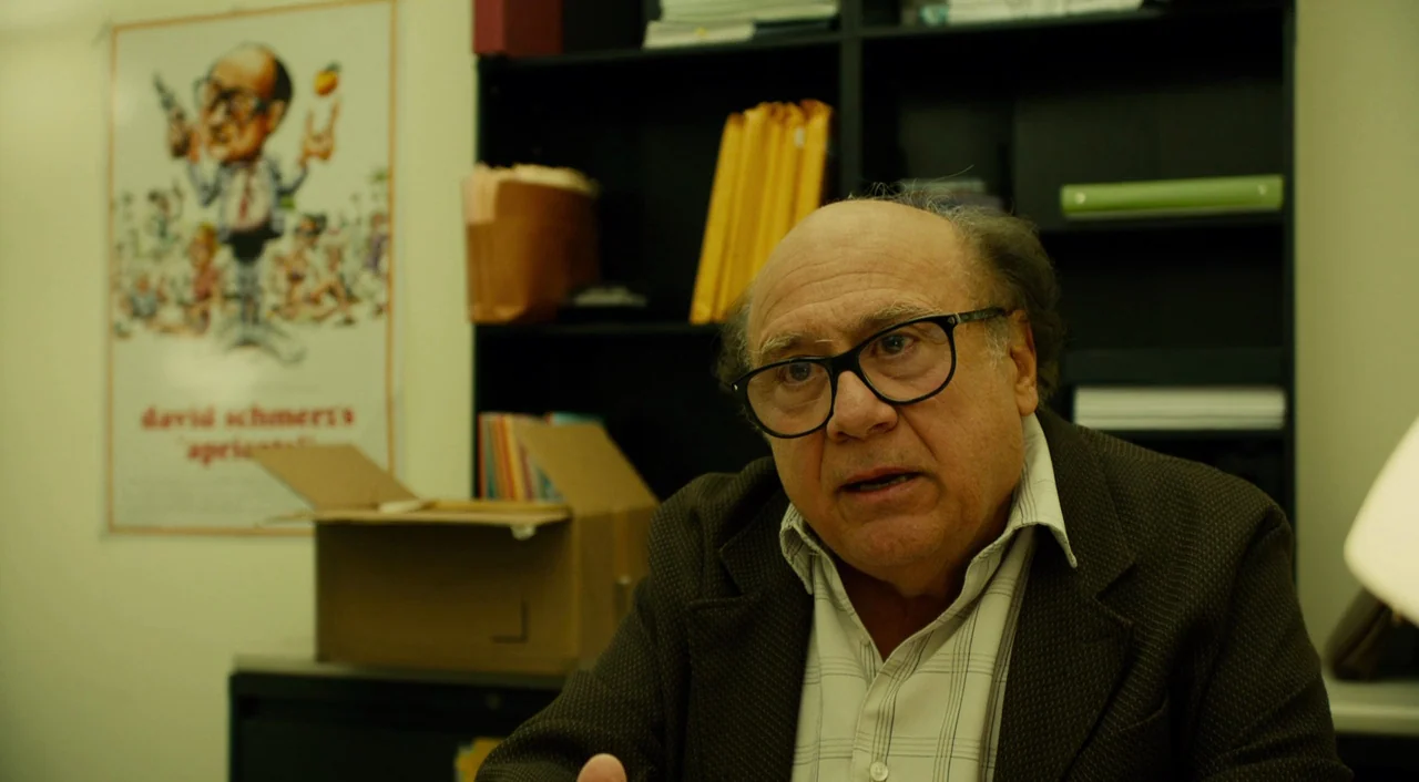 Wiener-Dog (2016) movie still: medium — An office space with a black bookshelf and a poster on the wall. An older man sits in front of the shelving…