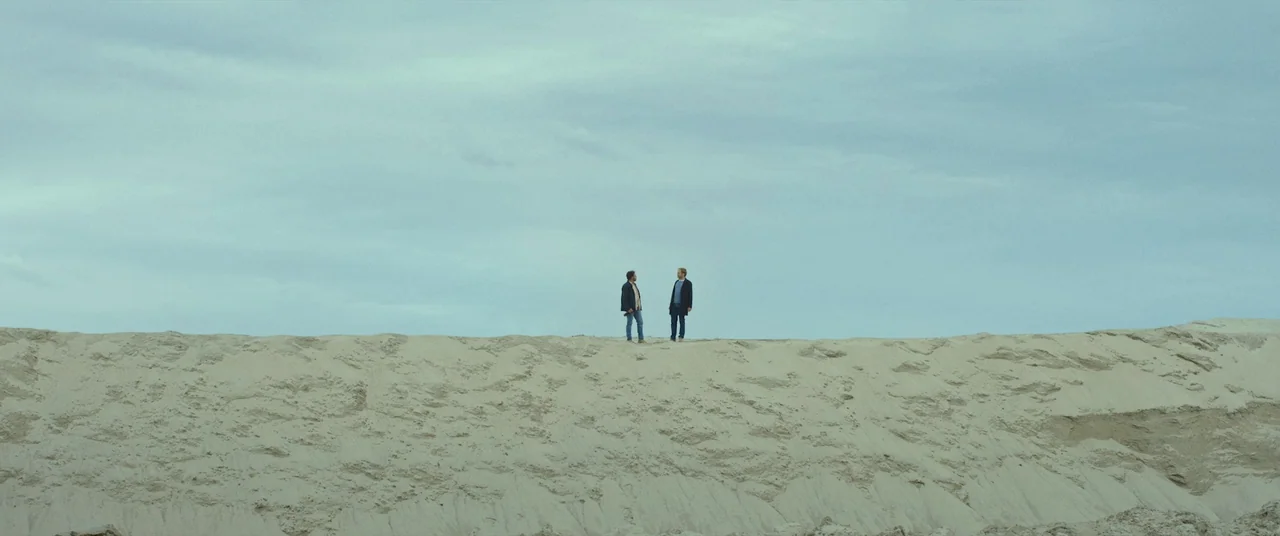 Speak No Evil (2022) movie still: wide — A vast expanse of tan sand dunes meets a flat, overcast grey-blue sky. Two men stand apart at the high…