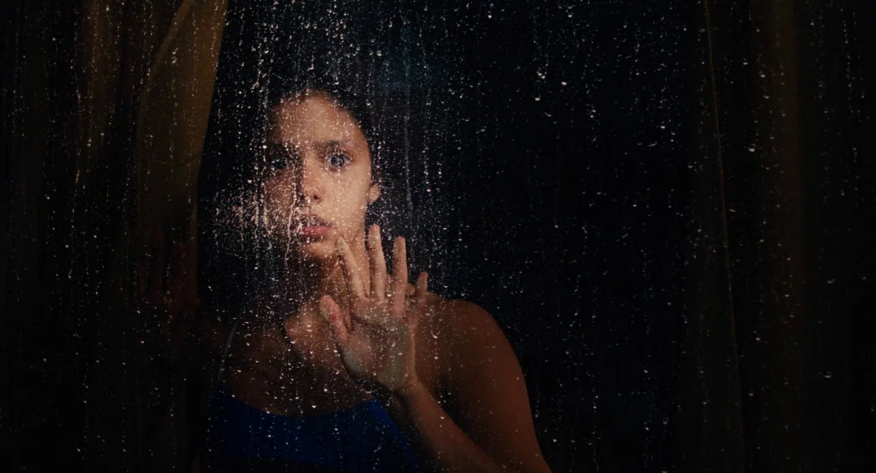 Ava (2017) movie still: closeup — A dark space seen through a wet, rain-covered glass window pane. A young woman stands behind the glass looking…