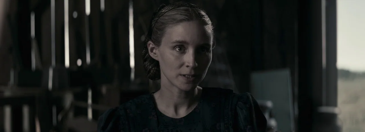 Women Talking (2022) movie still: medium — A woman stands in a dim rustic wooden structure with vertical planks and shadows. She is looking slightly…