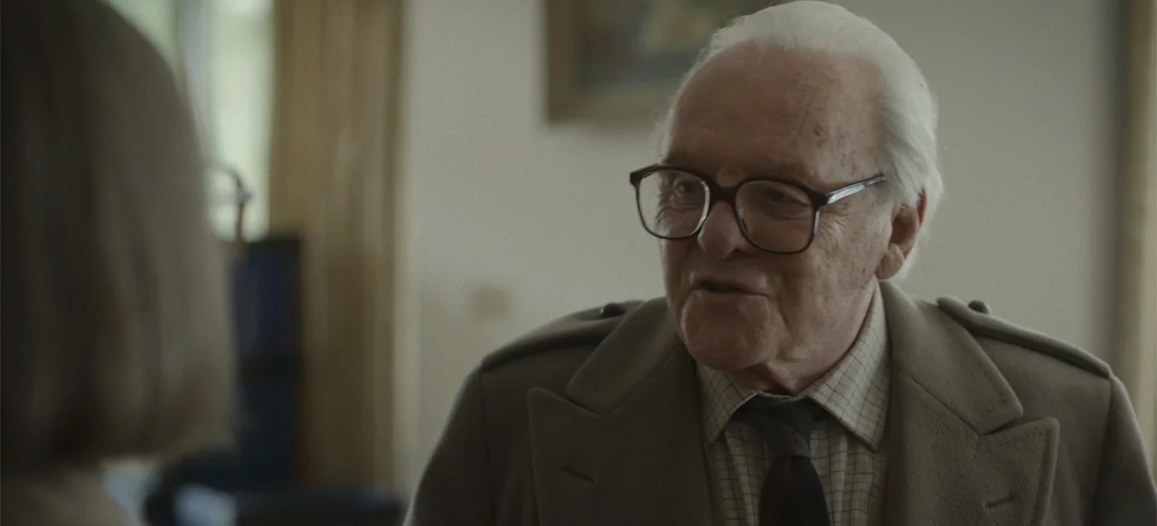 One Life (2023) movie still: medium — An interior space with pale walls and soft indoor lighting. An older man in a trench coat speaks while looking…