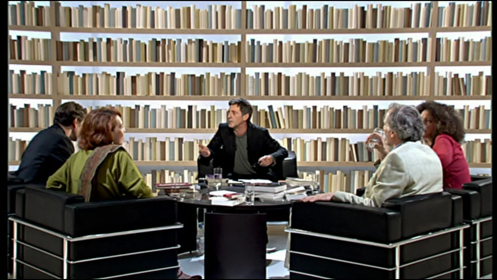 Caché (Hidden) (2005) movie still: wide — A television talk show set features five people seated around a table before bookshelves. They are…