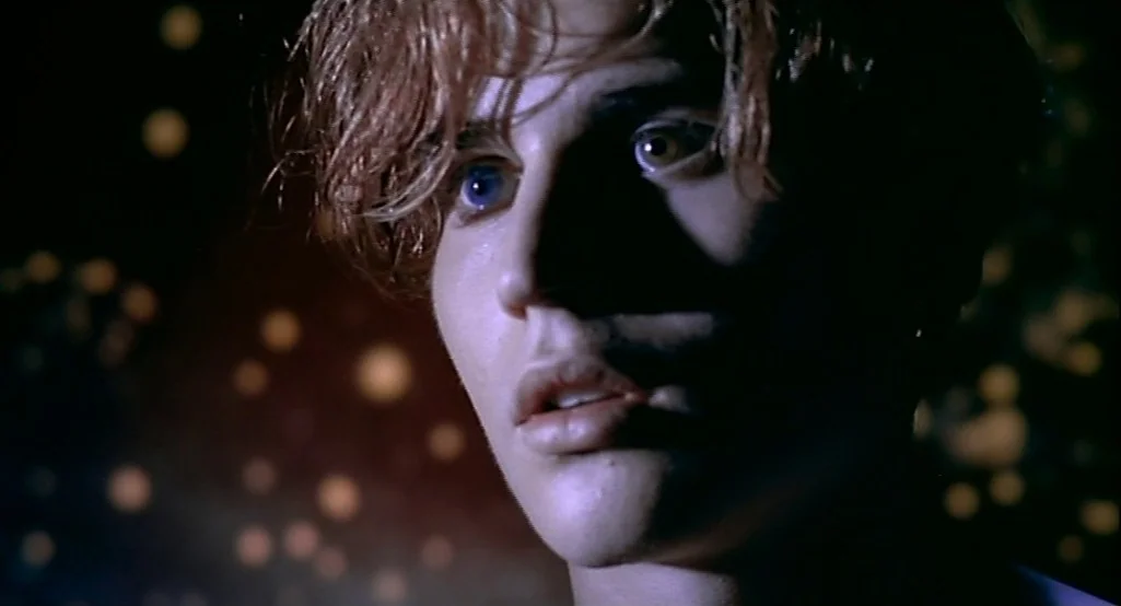 Nowhere (1997) movie still: closeup — A dark space features blurred yellow lights scattered against a deep black background. A young man with light…