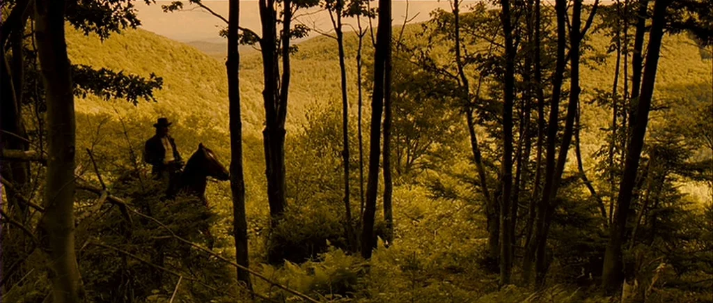 I’m Not There (2007) movie still: wide — A man on horseback stands on a steep, tree-covered hillside overlooking a valley. Tall trees frame the…