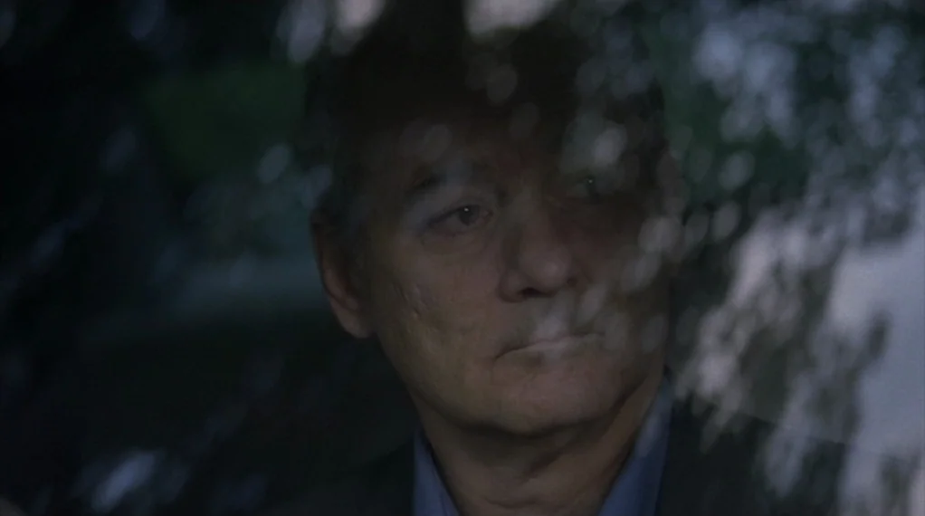 Broken Flowers movie still: closeup — An older man sits inside a vehicle looking through the side window. Reflections of tree foliage are visible…