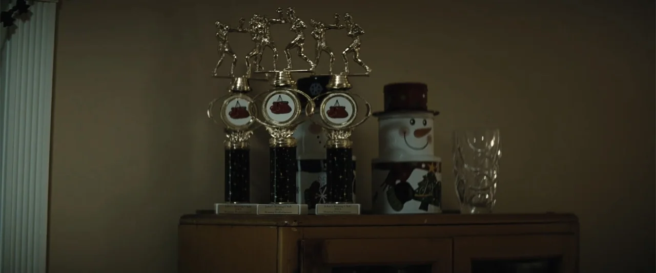 Run All Night (2015) movie still: medium — Three gold boxing trophies stand on a dark wooden shelf next to a snowman ornament. The background is a…