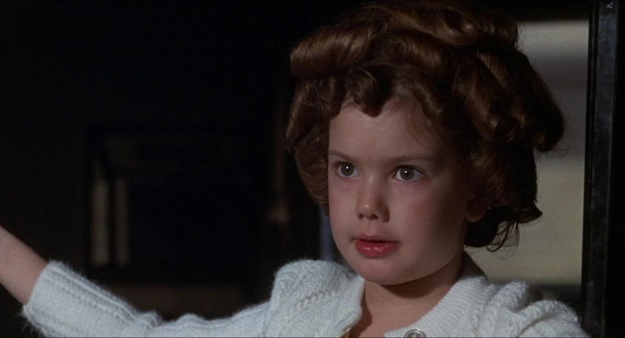 What’s the Matter with Helen? (1971) movie still: closeup — A dark indoor space with a young girl positioned in the foreground. The girl has curly…